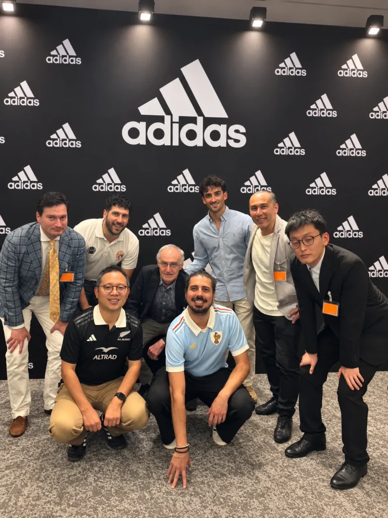 Eight attendees pose together in front of an adidas backdrop at a rugby community event in Tokyo.